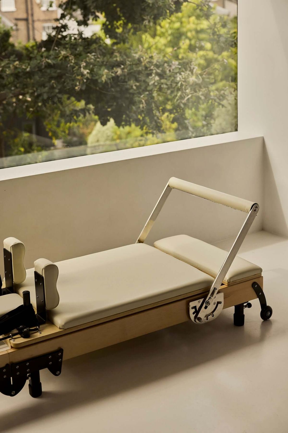 Cream-upholstered wooden Pilates reformer beside a large window overlooking trees