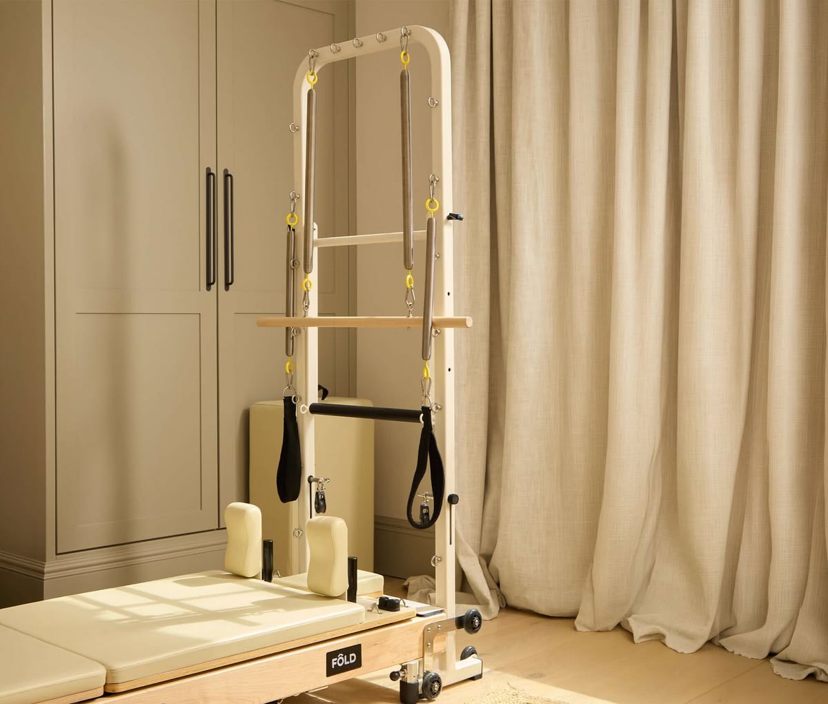 Wooden FÔLD Pilates reformer with cream upholstery and a tower beside fitted cupboards and linen curtains