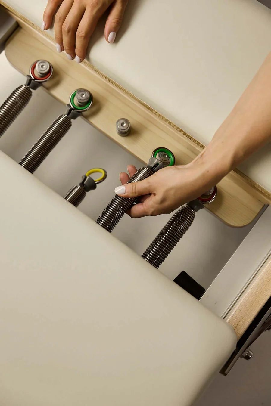 Hands adjusting colour-coded resistance springs beneath a cream-upholstered wooden Pilates reformer carriage
